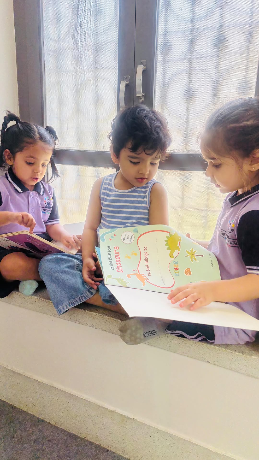 Three children sitting on a sunlit window ledge, turning the pages of a dinosaur sticker book together