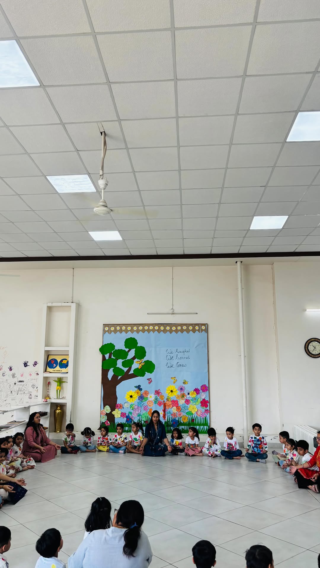 The whole school sitting cross-legged in a circle for morning assembly, under a painted board reading We laughed, we learned, we grew