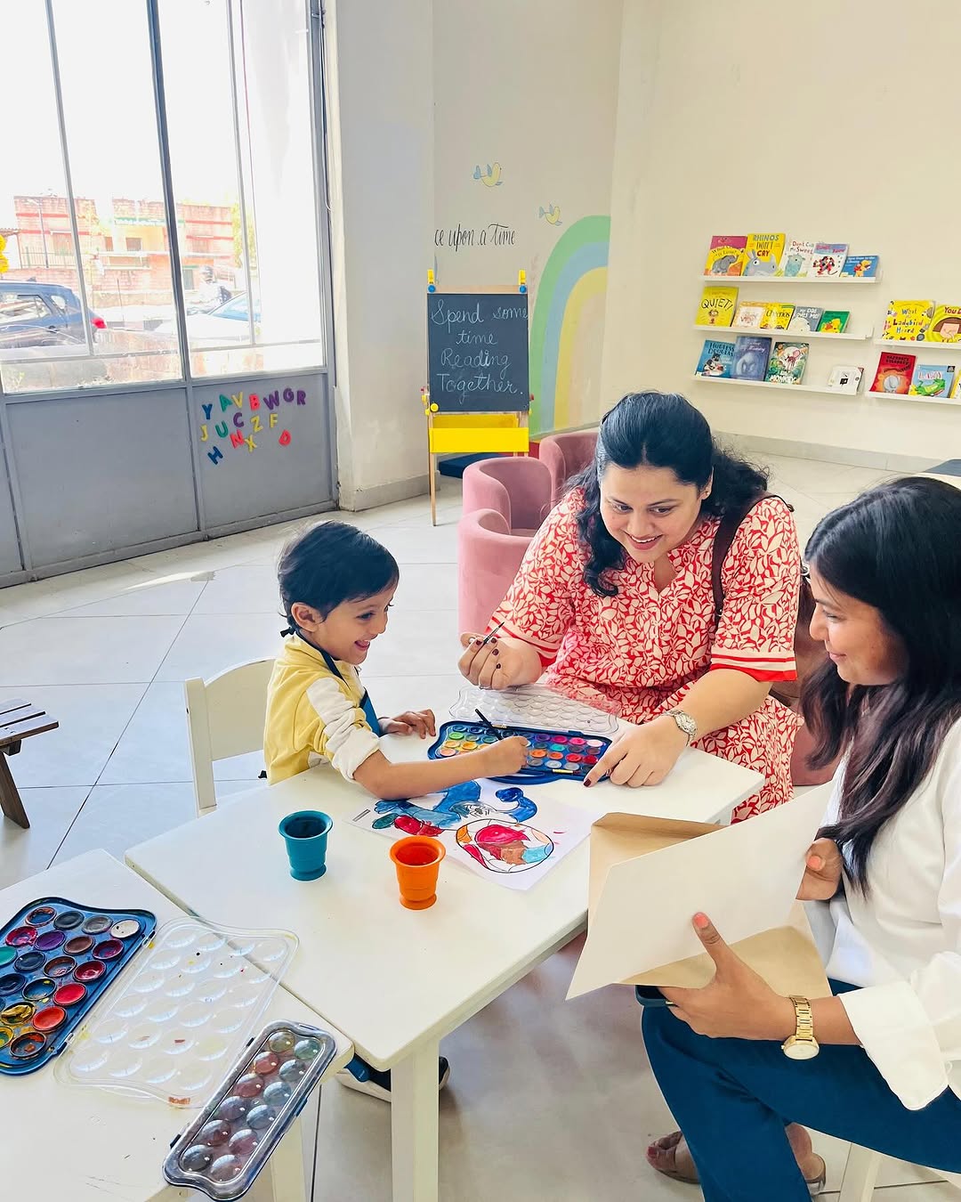 A child painting with watercolours at a white table while a parent and a teacher watch, beside the reading corner