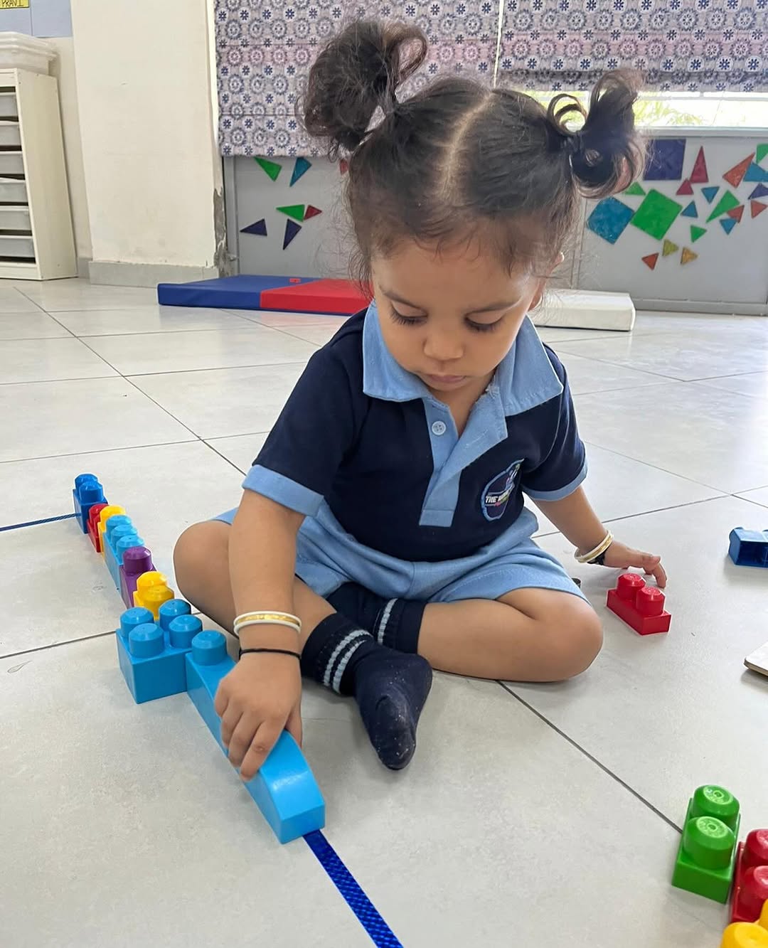 A toddler in school uniform building a long line of blocks on the classroom floor
